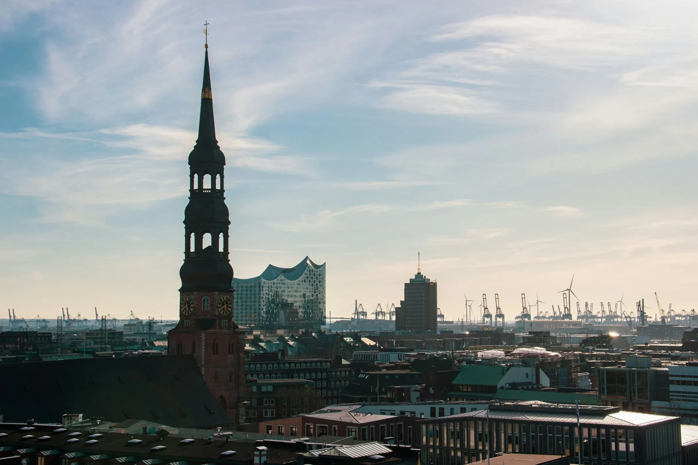 Hamburger Innenstadt-Panorama mit barockem Kirchturm im Vordergrund, dahinter Elbphilharmonie und die Kräne des Hamburger Hafens.